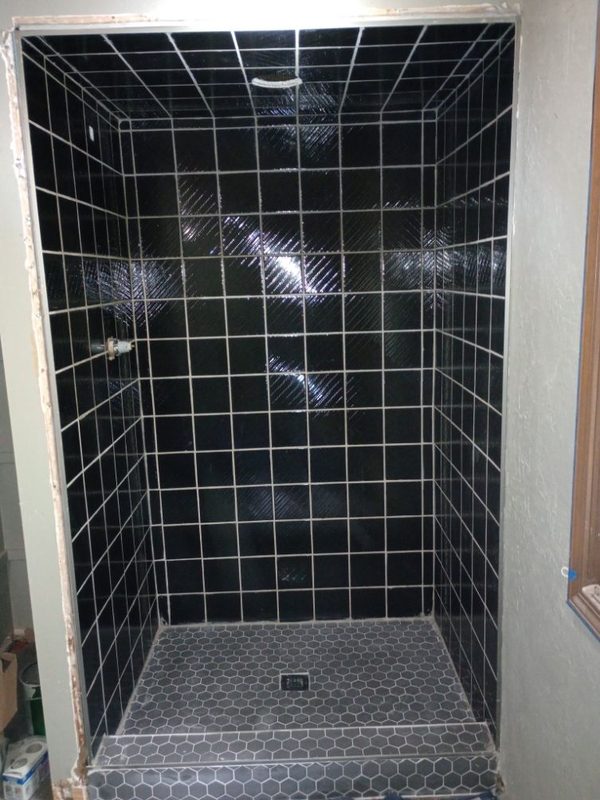 The same shower finished in glossy black square tile on the walls and ceiling, with a grey hexagon-tile floor and curb.