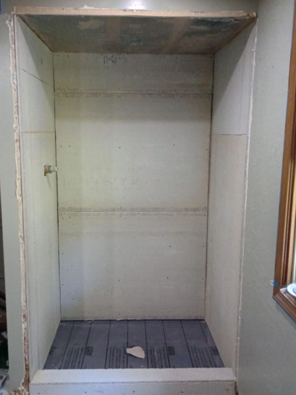 A walk-in shower enclosure with bare cement-board walls and ceiling, a membrane-covered floor and an unfinished curb, beside a window edged in blue painter's tape.