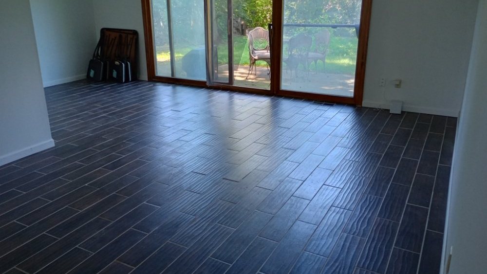 An empty room with a newly laid dark wood-look plank tile floor, lit by a sliding glass door that opens onto a patio and garden.
