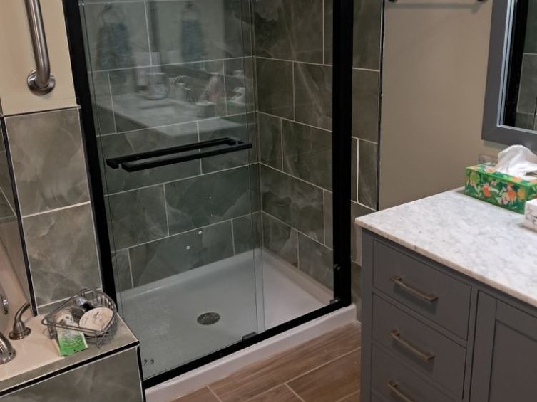 A bathroom with a black-framed glass shower door, grey marble-look wall tile, a grey vanity with a white marble top, and wood-look plank tile on the floor.
