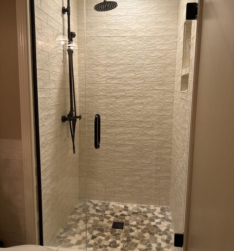 A narrow walk-in shower with textured white tile walls, a matte black rain shower head and handheld spray, a frameless glass door, and a floor of small river stones.